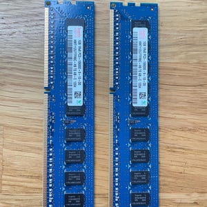 May include: Two blue computer RAM sticks with black chips and white text. Each stick has a label with the text "1GB 1Rx8 PC3-10600E-9-10-00" and other technical details. The sticks are on a wooden surface.