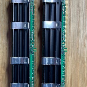 May include: Two computer RAM sticks. Each stick has a green circuit board with gold-colored pins and a black heat sink secured by silver metal clips. The components are arranged vertically.