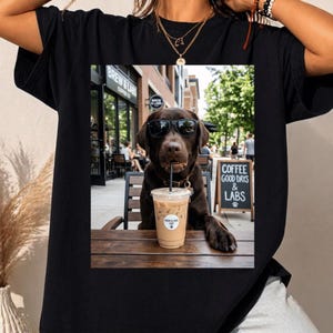 May include: A black t-shirt featuring a graphic print of a chocolate Labrador dog wearing black sunglasses, sitting at an outdoor cafe table with an iced coffee. The print includes text that reads "BREW & LABS" and "COFFEE GOOD DAYS & LABS".