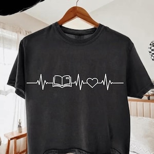 Peut inclure: Un t-shirt noir à manches courtes avec un imprimé graphique blanc représentant une ligne de rythme cardiaque, une icône de livre ouvert et un symbole de cœur centré sur la poitrine. Le t-shirt a un col rond et est sur un cintre en bois.