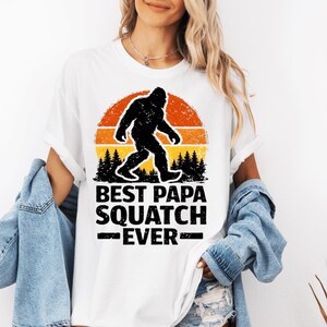 Peut inclure: T-shirt blanc avec une silhouette de Bigfoot noire sur un fond de coucher de soleil orange et jaune. Le texte "BEST PAPA SQUATCH EVER" est imprimé en noir. Un t-shirt décontracté et confortable.