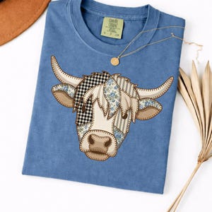 Peut inclure: Un t-shirt bleu avec un motif de tête de vache composé de divers empiècements à motifs, notamment du vichy noir et blanc, des fleurs bleues et des rayures beiges. Le t-shirt présente une encolure ronde et une étiquette au niveau du col.
