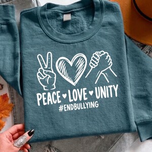 Op de afbeelding: Een donkergroenblauw sweatshirt met ronde hals en een witte grafische print van een vredesteken, een hart en twee in elkaar grijpende handen. Onder de afbeelding staat de tekst PEACE LOVE UNITY met hartjes en #ENDBULLYING.