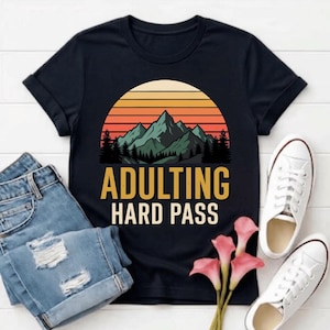 Peut inclure: Un t-shirt noir à manches courtes avec un motif de coucher de soleil sur une chaîne de montagnes et des pins. Sous le motif, le texte ADULTING HARD PASS est écrit en lettres jaunes et blanches. Le t-shirt est présenté avec un jean bleu.