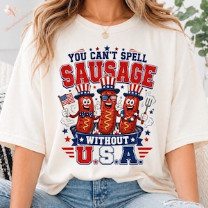 Peut inclure: T-shirt crème avec un graphisme indiquant "YOU CAN'T SPELL SAUSAGE WITHOUT U.S.A." Le graphisme présente trois hot-dogs de dessins animés portant des chapeaux et des lunettes de soleil patriotiques, avec un drapeau américain et une fourchette.