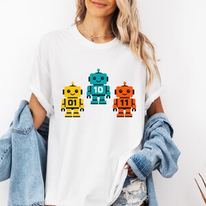 Peut inclure: T-shirt blanc avec trois robots colorés : jaune, turquoise et orange. Chaque robot porte un numéro : 01, 10 et 11. Les robots ont des détails noirs et se détachent sur un fond blanc. Vêtement décontracté.