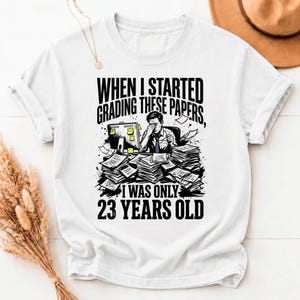 Puede incluir: Una camiseta blanca de manga corta con un gráfico de una persona sentada en un escritorio rodeada de pilas de papel. El texto dice: WHEN I STARTED GRADING THESE PAPERS, I WAS ONLY 23 YEARS OLD. El diseño incluye un monitor de computadora.