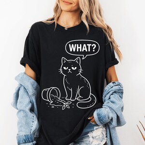 Peut inclure: T-shirt noir avec une illustration d'un chat noir grincheux et le mot "WHAT?" dans une bulle de dialogue. Le chat est à côté d'un pot renversé. Design humoristique et décontracté.