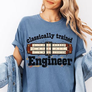 Puede incluir: Una camiseta azul con un gráfico de una regla de cálculo vintage con escalas numéricas. El texto "classically trained" aparece en negro sobre la regla y la palabra "Engineer" está impresa en letras negras grandes debajo de ella.