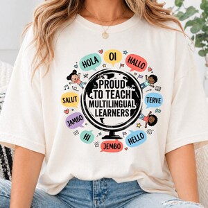 May include: An off-white t-shirt featuring a central black globe graphic with the text PROUD TO TEACH MULTILINGUAL LEARNERS. Surrounding the globe are colorful speech bubbles containing greetings in various languages and illustrations of children.