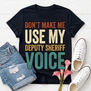 Peut inclure: Un t-shirt noir à manches courtes avec le texte DON'T MAKE ME USE MY DEPUTY SHERIFF VOICE dans une police de style rétro aux couleurs orange, crème et sarcelle. Le t-shirt est présenté avec un jean bleu et des baskets blanches.