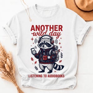 Peut inclure: Un t-shirt blanc avec une illustration de raton laveur portant un sweat à capuche, un casque et un sac à dos. Le raton laveur tient un smartphone et une tasse rouge marquée WILD. Le texte indique ANOTHER wild day LISTENING TO AUDIOBOOKS.