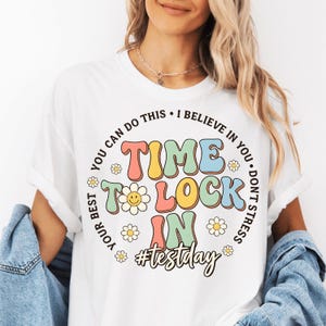 Peut inclure: T-shirt blanc avec un graphisme coloré indiquant "Time to Lock In" avec un hashtag #testday. Le motif comprend une marguerite et les phrases "You can do this", "I believe in you", "Don't stress" et "Your best".