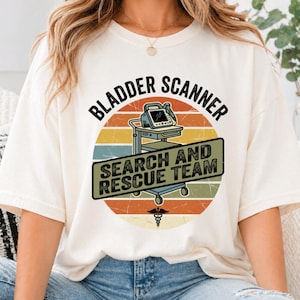 Puede incluir: Una camiseta color crema con un gráfico de un escáner de vejiga médico sobre un carrito. El texto dice BLADDER SCANNER SEARCH AND RESCUE TEAM. El diseño incluye un fondo de atardecer retro en naranja, amarillo y azul, con un símbolo de caduceo.
