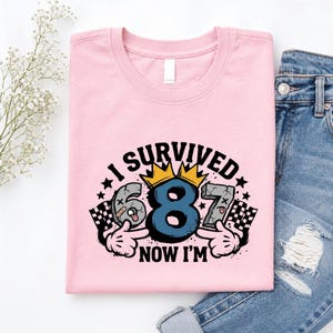 Könnte beinhalten: Ein hellrosa T-Shirt mit einem Aufdruck, der den Text I SURVIVED 687 NOW I'M zeigt. Das Design enthält eine große blaue 8 mit einer gelben Krone, flankiert von einer grauen 6 und 7, Cartoon-Händen sowie schwarz-weißen Zielflaggen.