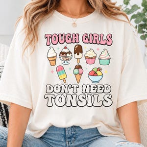 May include: An off-white t-shirt featuring a graphic of various ice cream treats and the text "TOUGH GIRLS DON'T NEED TONSILS" in pink and black lettering. The design includes illustrations of soft serve, sundaes, ice lollies, and cupcakes.