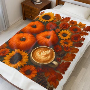 Könnte beinhalten: Eine Decke mit einem herbstlichen Design. Das Design zeigt orangefarbene Kürbisse, Sonnenblumen und rote und orangefarbene Blätter. Eine Tasse Kaffee mit Latte Art ist ebenfalls abgebildet. Die Decke liegt auf einem Bett.