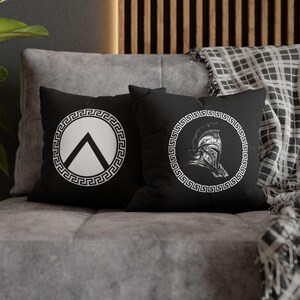 May include: Two square black throw pillows featuring white graphic prints. One pillow displays a circular Greek key border surrounding a Spartan shield symbol. The other pillow features a circular Greek key border enclosing a detailed Spartan helmet.