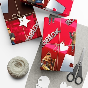 May include: Two rectangular gifts wrapped in bright red paper featuring the Roblox logo and various character graphics. Each package is tied with brown twine and adorned with a white paper gift tag in the shape of a star or a heart.