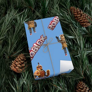 Puede incluir: Un regalo rectangular envuelto en papel azul claro con un patrón repetitivo de personajes de Roblox y el logotipo rojo de Roblox. El paquete está atado con un cordel marrón fino y una esquina del papel de regalo está ligeramente doblada.
