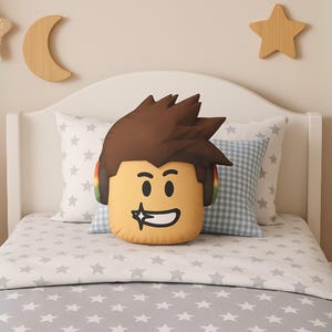 May include: A plush throw pillow shaped like a stylized character head with brown spiky hair, a tan complexion, and a wide grin featuring a sparkling tooth. The character wears colorful headphones over the ears, with a smooth, soft fabric finish.