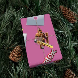 Puede incluir: Una caja de regalo rectangular envuelta en papel rosa con un patrón repetitivo de un caballero con armadura dorada que sostiene una espada y un escudo, junto con el logotipo de Roblox, atada con un cordel fino de color marrón.