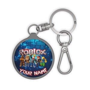 May include: A silver keychain with a round, blue and silver charm featuring a group of Roblox characters and the text "Roblox" in red. The charm also has the text "Your Name" in white.