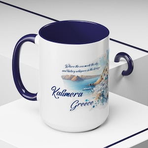 Peut inclure: Mug en céramique blanche avec une bordure et une anse bleues. Le mug présente une illustration aquarelle d'une île grecque avec le texte "Where the sea meets the sky and history whispers in the breeze" et "Kalimera Greece".