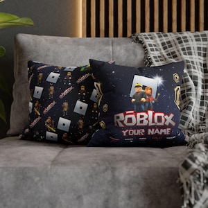 May include: Two decorative throw pillows with a Roblox theme. One pillow features a repeating pattern of Roblox characters and logos on a black background. The other pillow features a Roblox character with the text "Roblox Your Name" on a black background with a starry sky design.
