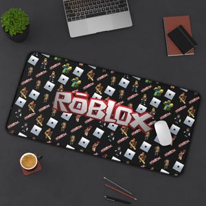 May include: Black rectangular mousepad featuring a repeating pattern of Roblox characters and logos. The large Roblox logo is prominently displayed in the center. A computer mouse rests on the pad, alongside a laptop, coffee cup, and other office supplies.