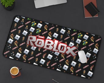 May include: A rectangular desk mat featuring a black background with a repeating pattern of small Roblox character figures, white square icons, and the word Roblox. A large, centered, silver and red Roblox logo is prominently displayed on the mat.