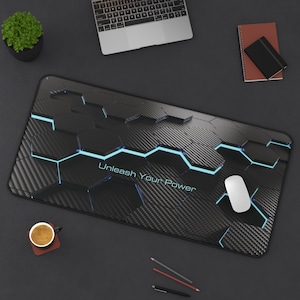 May include: A rectangular desk mat featuring a black carbon fibre texture with a geometric hexagonal pattern and glowing blue light accents. The text Unleash Your Power is printed in the centre in a light blue font, with a white computer mouse.