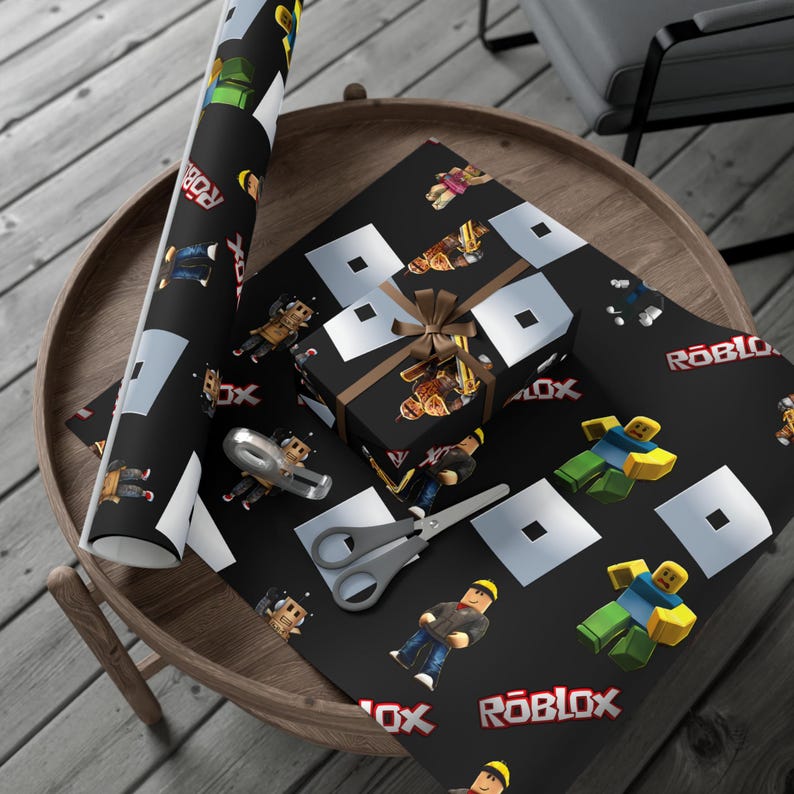 May include: A roll of black wrapping paper and a gift box wrapped in the same paper, both featuring a repeating pattern of Roblox characters, the Roblox logo, and white square icons. A pair of silver scissors rests on the flat sheet of paper.
