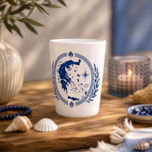 Könnte beinhalten: Ein weißer Keramikbecher mit einem blauen Grafikdruck der Landkarte von Griechenland in einem ovalen Rahmen. Das Design umfasst eine Windrose, stilisierte Lorbeerzweige und ein traditionelles griechisches Mäander-Muster als Rand.