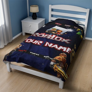 May include: A dark blue blanket featuring a space-themed background with Roblox character graphics, the Roblox logo, and the text "YOUR NAME" in large white letters. The design includes multiple black horizontal bands with the Roblox logo and years.