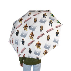 Könnte beinhalten: Ein weißer Regenschirm mit einem sich wiederholenden Muster aus verschiedenen Roblox-Charakterfiguren, dem roten Roblox-Logo und hellgrauen quadratischen Symbolen. Das Design zeigt diverse Avatare in verschiedenen Outfits und Posen.