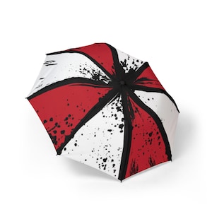 Könnte beinhalten: Ein faltbarer Regenschirm mit einem radialen Muster aus abwechselnd roten und weißen Feldern. Das Design zeigt schwarze, tintenartige Spritzer und Pinselstrich-Texturen, die von der Mitte ausgehen, sowie dicke schwarze Linien zwischen den Feldern.