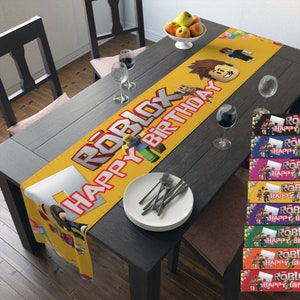 May include: A yellow table runner with the words "Roblox Happy Birthday" printed in red and black. The runner features a cartoon Roblox character and balloons. There are also several smaller banners with the same design in different colors.