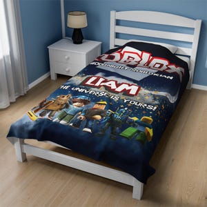 Op de afbeelding: Een blauwe en zwarte fleece deken in tweepersoonsformaat met een Roblox-logo, de tekst "Goodnight Robloxian", een naam "Liam" en de tekst "The Universe is Yours!" De deken heeft een cartoon afbeelding van Roblox-karakters.