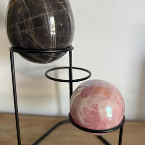 May include: A black metal tiered stand holding a large, dark brown, egg-shaped stone and a smaller, pink sphere. The egg-shaped stone has a patterned surface. The sphere has iridescent highlights. The stand is on a wooden surface.