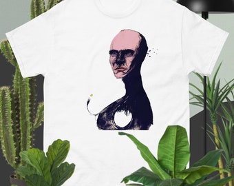 Può includere: Maglietta bianca con un'illustrazione in bianco e nero di una persona con il viso rosa e un buco nel petto.