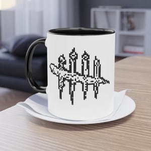 Könnte beinhalten: Eine weiße Keramik-Tasse mit schwarzem Innenraum und Henkel. Die Tasse zeigt eine schwarz-weiße pixelige Grafik eines stilisierten Objekts. Die Tasse steht auf einer weißen Untertasse auf einer Holzoberfläche.