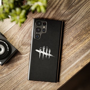 Puede incluir: Funda negra para smartphone con un dise&ntilde;o blanco de marca de conteo tachada. La funda est&aacute; sobre una superficie de madera. Una c&aacute;mara y vegetaci&oacute;n son visibles en el fondo. La funda tiene una textura.