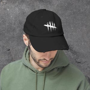 Puede incluir: Gorra de béisbol negra con visera desgastada. La gorra presenta un gráfico blanco de cinco líneas verticales con una línea horizontal que las atraviesa. La persona lleva una sudadera con capucha verde.