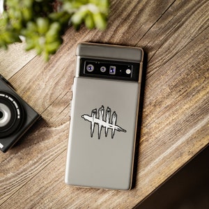Puede incluir: Una funda gris para smartphone con un gr&aacute;fico en blanco y negro de un s&iacute;mbolo cruzado. La funda del tel&eacute;fono est&aacute; sobre una superficie de madera, con una c&aacute;mara y vegetaci&oacute;n en el fondo.