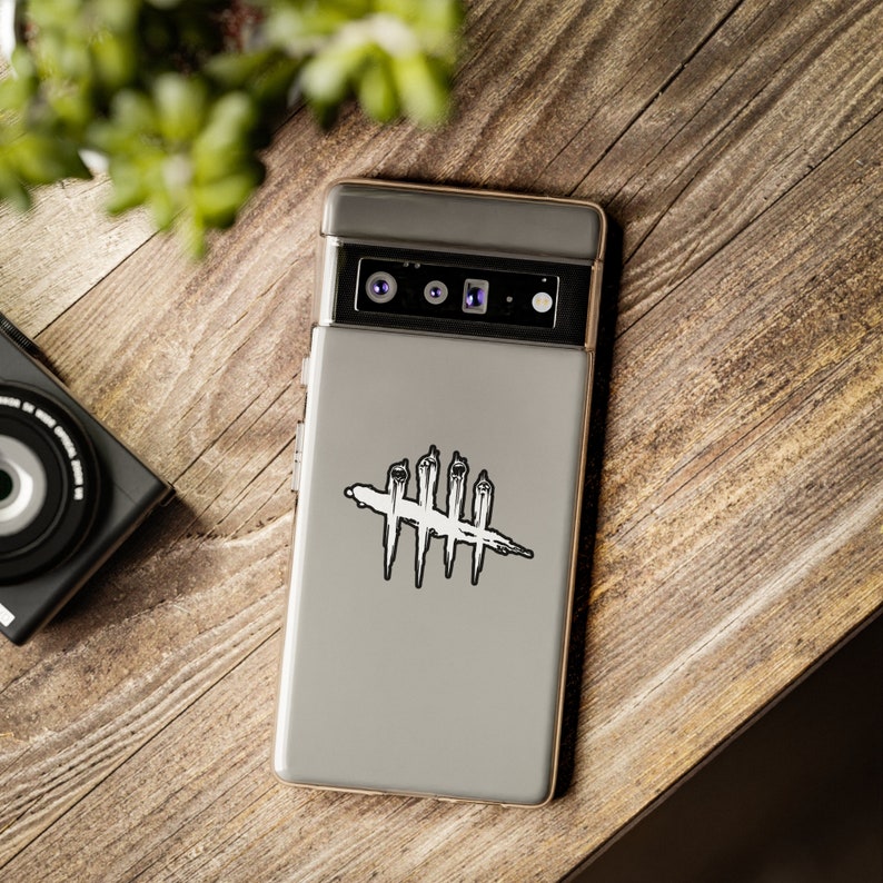 Puede incluir: Una funda gris para smartphone con un gr&aacute;fico en blanco y negro de un s&iacute;mbolo cruzado. La funda del tel&eacute;fono est&aacute; sobre una superficie de madera, con una c&aacute;mara y vegetaci&oacute;n en el fondo.