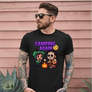 May include: Black t-shirt with a graphic of two cartoon characters camping. One character is wearing a green hooded outfit and crying. The other character is holding a marshmallow over a campfire. The text "CAMPING AGAIN" is above the characters.