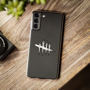 Puede incluir: Un smartphone negro con una funda transparente, con un dise&ntilde;o gr&aacute;fico en blanco y negro. El dise&ntilde;o es un s&iacute;mbolo estilizado con l&iacute;neas verticales cruzadas por una l&iacute;nea horizontal. El tel&eacute;fono est&aacute; sobre una superficie de madera, con una c&aacute;mara visible en el fondo.