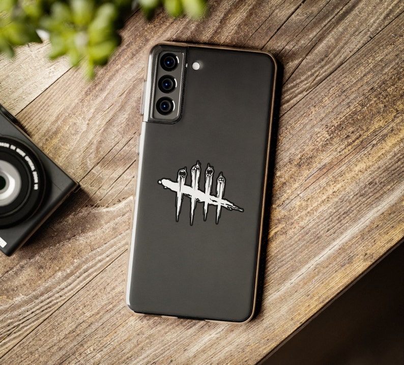 Puede incluir: Un smartphone negro con una funda transparente, con un dise&ntilde;o gr&aacute;fico en blanco y negro. El dise&ntilde;o es un s&iacute;mbolo estilizado con l&iacute;neas verticales cruzadas por una l&iacute;nea horizontal. El tel&eacute;fono est&aacute; sobre una superficie de madera, con una c&aacute;mara visible en el fondo.