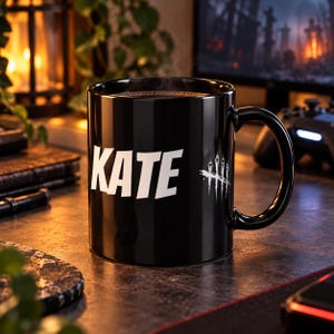 Può includere: Una tazza in ceramica nera con il testo bianco KATE stampato sul davanti, accompagnato da un simbolo grafico bianco composto da quattro linee verticali sormontate da una piccola forma a diamante, posizionato a destra del nome sulla tazza.